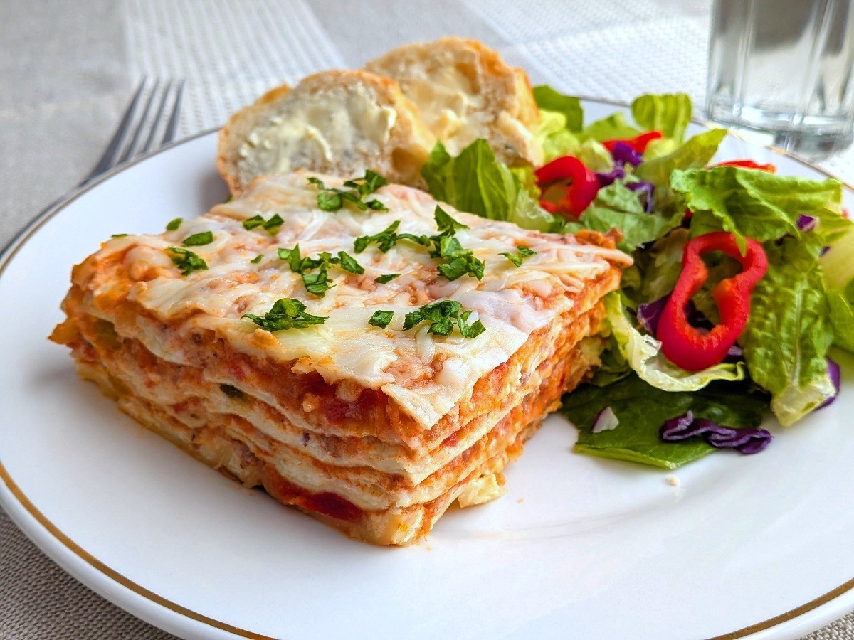 A slice of lasagna on a plate with buttered bread and salad.
