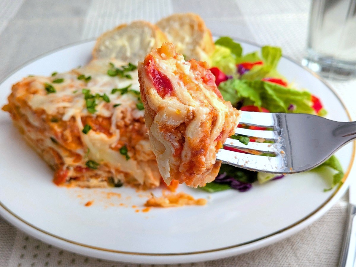 A bit of lasagna on a fork. The rest of the lasagna serving can be seen on a white plate in the background with buttered bread and green salad.