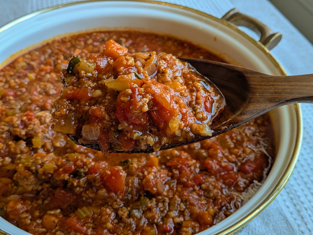 A close up of a large serving spoon full of pasta sauce with visible tomatoes, zucchini, and carrots.