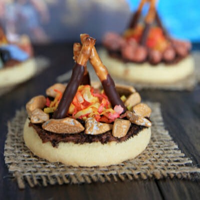 A cute shortbread or sugar cookie, topped with chocolate frosting, chocolate rocks, dyed coconut, and chocolate-dipped mini pretzel rods, made to look like a little fire pit.