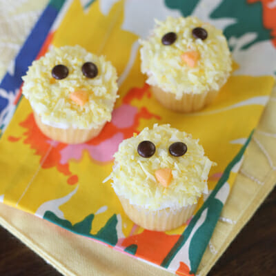 Mini cupcakes that are frosted and sprinkled with pale yellow coconut flakes. They are decorated to look like chicks with two mini brown M&Ms for eyes and a small beak made out of an orange Starburst candy.