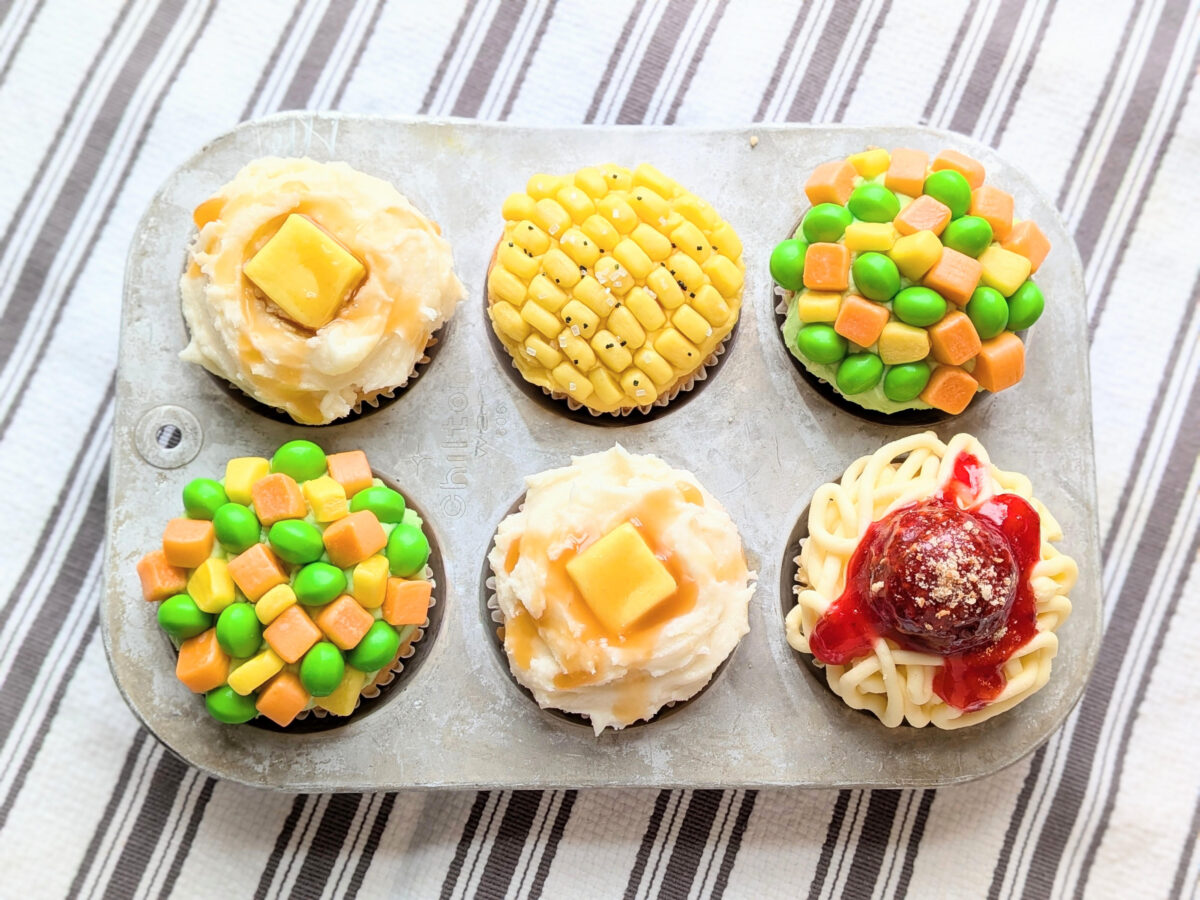 A pan of silly cupcakes, decorated to look like mixed veggies, mashed potatoes with gravy, and spaghetti and meatballs.