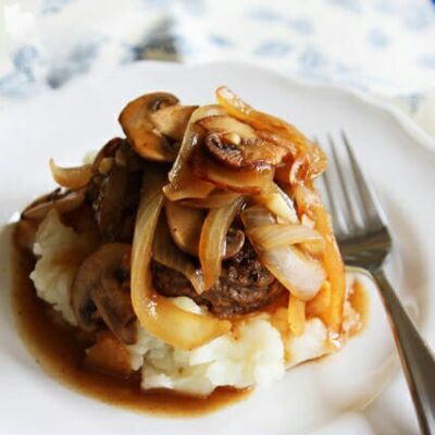 A pile of mashed potatoes, topped with a hamburger steak smothered in mushrooms and onions.