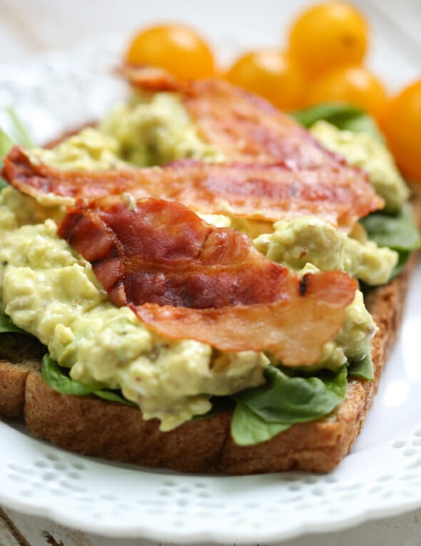 A piece of toast topped with avocado egg salad and slices of bacon. 