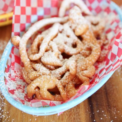 A funnel cake, dusted in powdered sugar, sitting in a blue basket lined with red checkered parchment.