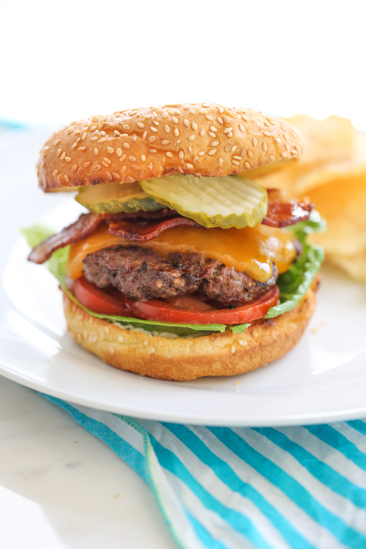 Cheeseburger with toppings on a while plate with chips in the background