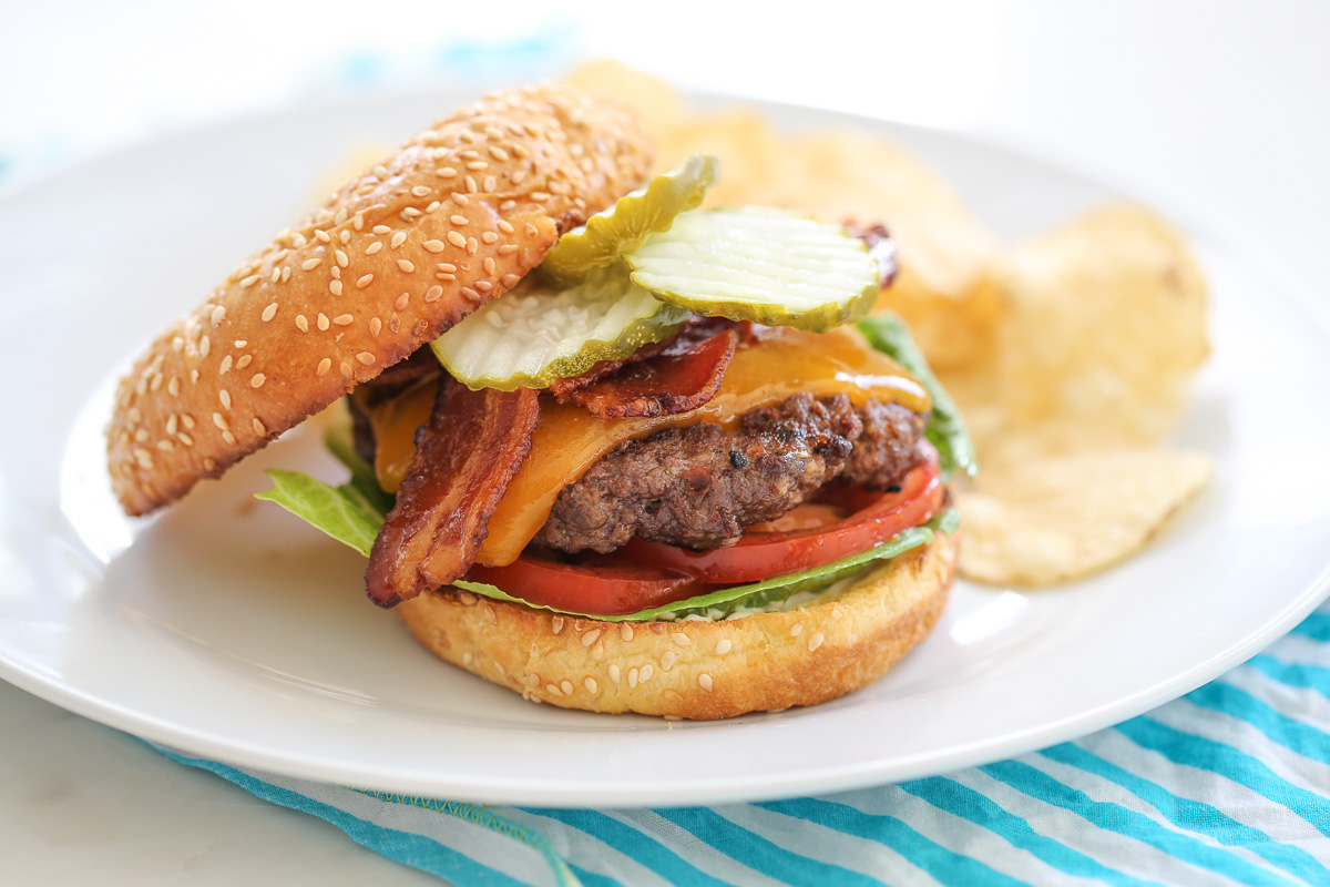 Cheeseburger with toppings on a while plate with chips in the background