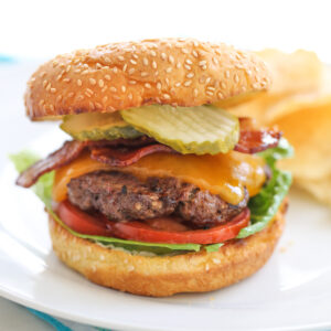 Cheeseburger with toppings on a while plate with chips in the background