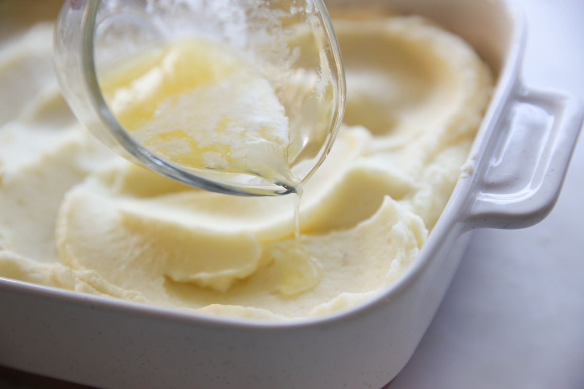 pouring butter over creamy mashed potatoes in a white bowl
