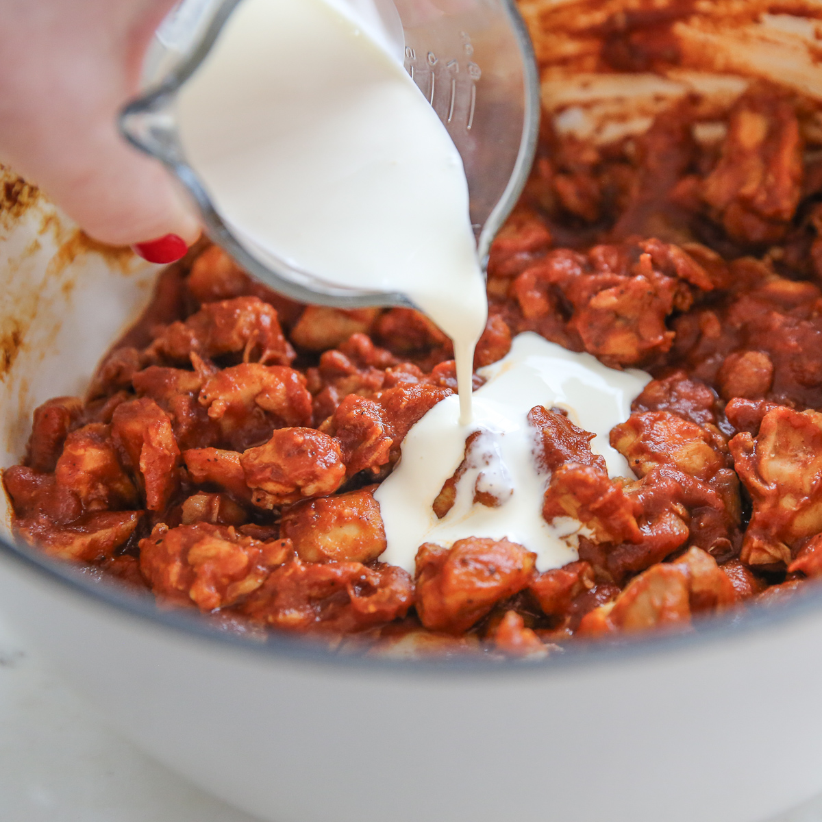 pouring cream into a pot of chicken cooked with tomato and seasonings