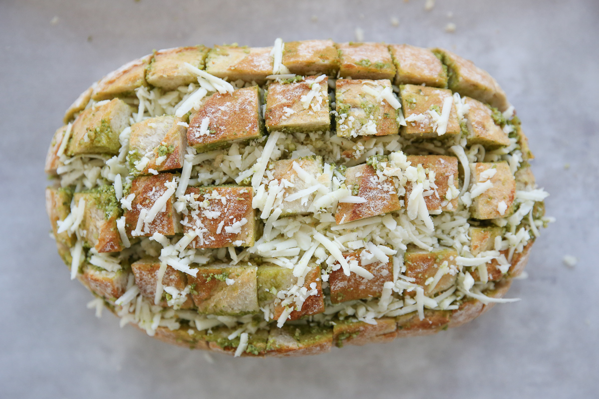 preparing a loaf of bread with pesto and shredded cheese